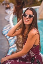 A young brunette in a maroon floral dress and trendy white sunglasses enjoying the summer in the golden hour at a city fountain