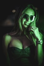 Caucasian brunette in a black dress illuminated by green led light reflected in black crystals. Urban night photography. Girl with sunglasses and saying quiet