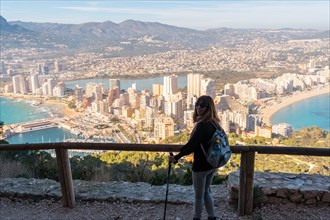 A young hiker girl with a hat looking at the city of Calpe from the path of the Penon de Ifach Natural Park