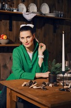 Kind woman in warm green knitted sweater sitting behind dining table with candle on it in the kitchen of her wooden cabin in the evening