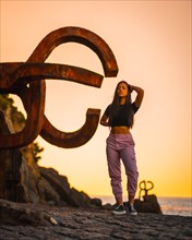 A young and pretty brunette Latina with long straight hair in a short black T-shirt and pink pants. Next to a sculpture of San Sebatian called Peine del Viento at sunset