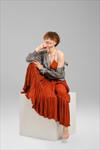 Smiling barefoot woman in sundress and shirt sits on cube in bright studio