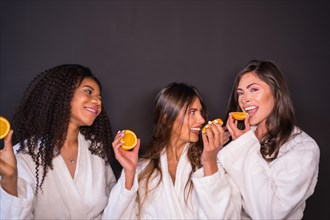 Studio photo with grey background of multi-ethnic relaxed women in bathrobe eating fruit
