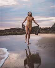 A brunette woman having fun with a swimsuit and a pareo walking along the beach in summer in the cloudy sunset