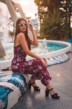 Portrait of a young brunette in a maroon floral dress and modern white sunglasses enjoying the summer in the golden hour in a city fountain