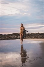 A brunette woman with a swimsuit and a pareo walking along the beach in summer in the cloudy sunset