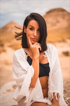 Sensual look of a young brunette Caucasian girl in a white dress sitting in a beautiful desert