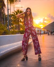 Sunset with a young brunette in a maroon floral dress and modern white sunglasses enjoying the summer in the golden hour