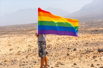 A gay person in a gray t-shirt and black cap with the LGBT flag waving in a desert with the wind