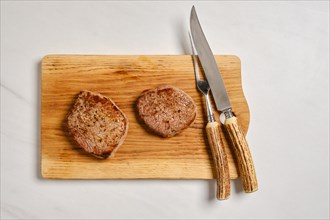 Top view of juicy beef steak on wooden cutting board next to fork and knife