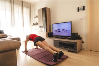 A young woman doing push-ups in the room watching TV. Sport in the covid19 quarantine at home