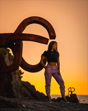 A young pretty brunette Latina with long straight hair in a short black T-shirt and pink pants. Orange sunset next to a famous sculpture of San Sebatian called Peine del Viento