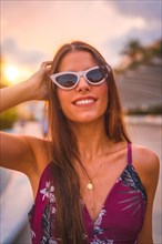 Portrait at sunset with a brunette in a maroon floral dress and white sunglasses enjoying the summer in the golden hour