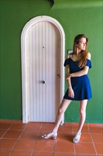 Lifestyle of a young caucasian girl in a blue dress in a summer perched next to a white door