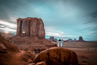 A boy in a white T-shirt sitting on a stone with his arms up in the sunset inside Monument Valley National Park. Utah