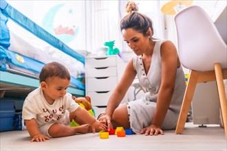 Young Caucasian mother playing with her in the room with toys. Baby less than a year learning the first lessons of her mother. Mother playing with her son playing sitting on the floor