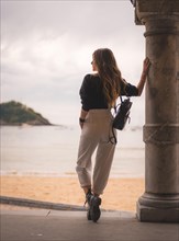 Lifestyle in the city with a blonde girl in white pants and a leather jacket near the beach. Photos next to a column looking at the beautiful beach