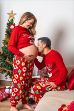 Young couple with decoration and red Christmas clothes kissing pregnant girlfriend's belly. Family with pregnant woman