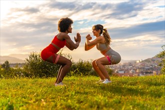 Caucasian blonde girl and dark-skinned girl with afro hair doing squat exercises in a park with the city in the background. Healthy life