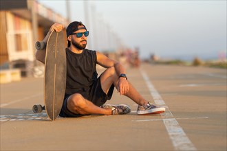A cool guy sitting on a road with a skateboard. Mid shot
