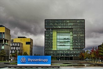 Dark clouds gather over in the background headquarters of steel giant company of steel production manufacturer of steel thyssenkrupp Thyssen Krupp in the foreground logo lettering