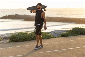 A man walking on the road near by beach holding a skateboard on his shoulder. Mid shot