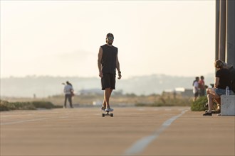 Adult attractive smiling man in black clothes skating on the street by the beach. Mid shot