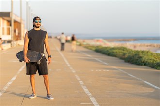 A man standing on the bike path holding a skateboard. Mid shot