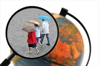 Tourists walking in the rain with umbrellas in summer seen through magnifying glass held against illuminated terrestrial globe