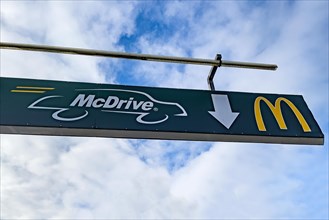 Sign mounted at height on pole with crossbar with white lettering McDrive and logo yellow M of McDonald's above entrance from drive in to fast food restaurant