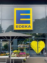 Logo with letter and lettering above entrance to supermarket below note on opening hours Monday to Saturday 7.00 a.m. to 9.00 p.m. to the right stylised heart of retail chain supermarket chain Edeka f...