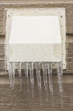 Metal exhaust vent covered with ice and icicles on tan clapboard wall of house