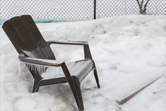Brown plastic Adirondack armchair covered in ice in backyard after freezing rainstorm in early spring