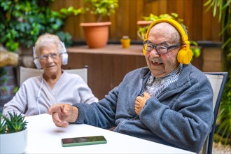 Happy old man listening to music with headphones in a geriatric next to other elder people