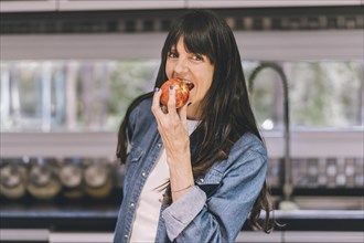 Woman eating an apple while looking at camera in her kitchen