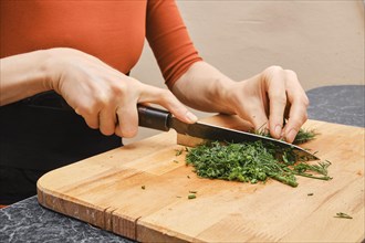 Unrecognizable woman cutting fresh dill on cutting board while sitting behind kitchen table