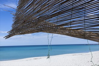 Straw mat hanging in the wind on a hut on a white sandy beach in front of a bright blue sea
