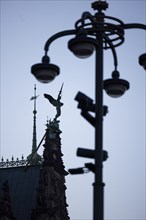 Angel figure with sword on the roof of the town hall fighting against a pole with lamps and video cameras