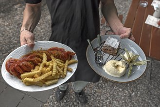 A waiter serves currywurst with fries and apple strudel with ice cream in a garden restaurant