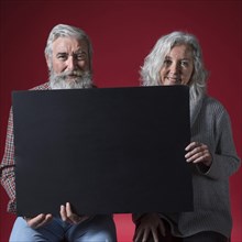 Portrait happy senior couple holding black placard looking camera against red background