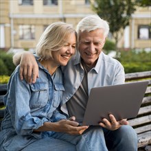 Smiley older couple sitting bench outdoors with laptop