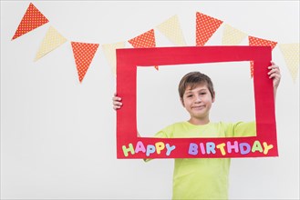 Smiling boy holding frame with happy birthday frame against wall decorated with bunting