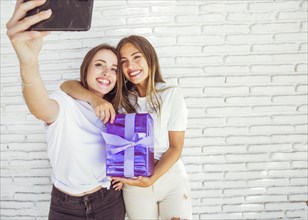 Two smiling female friends with gift taking selfie smartphone