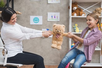Female psychologist smiling girl sitting face face pulling teddybear