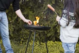 Girl with sausage standing near her father grilling portable barbecue