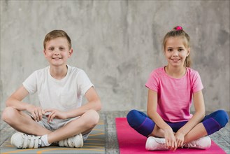 Portrait smiling girl boy sitting exercise mat with their crossed legs front wall