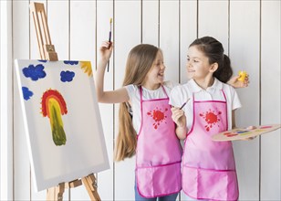Portrait smiling two girls pink apron making fun while painting canvas