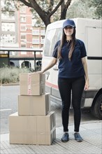 Portrait young female courier standing with stacked cardboard boxes