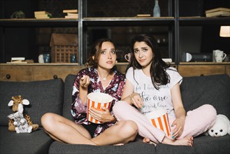 Two scared women sitting sofa with popcorn watching television