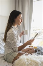 Young woman sitting bed reading newspaper while drinking coffee